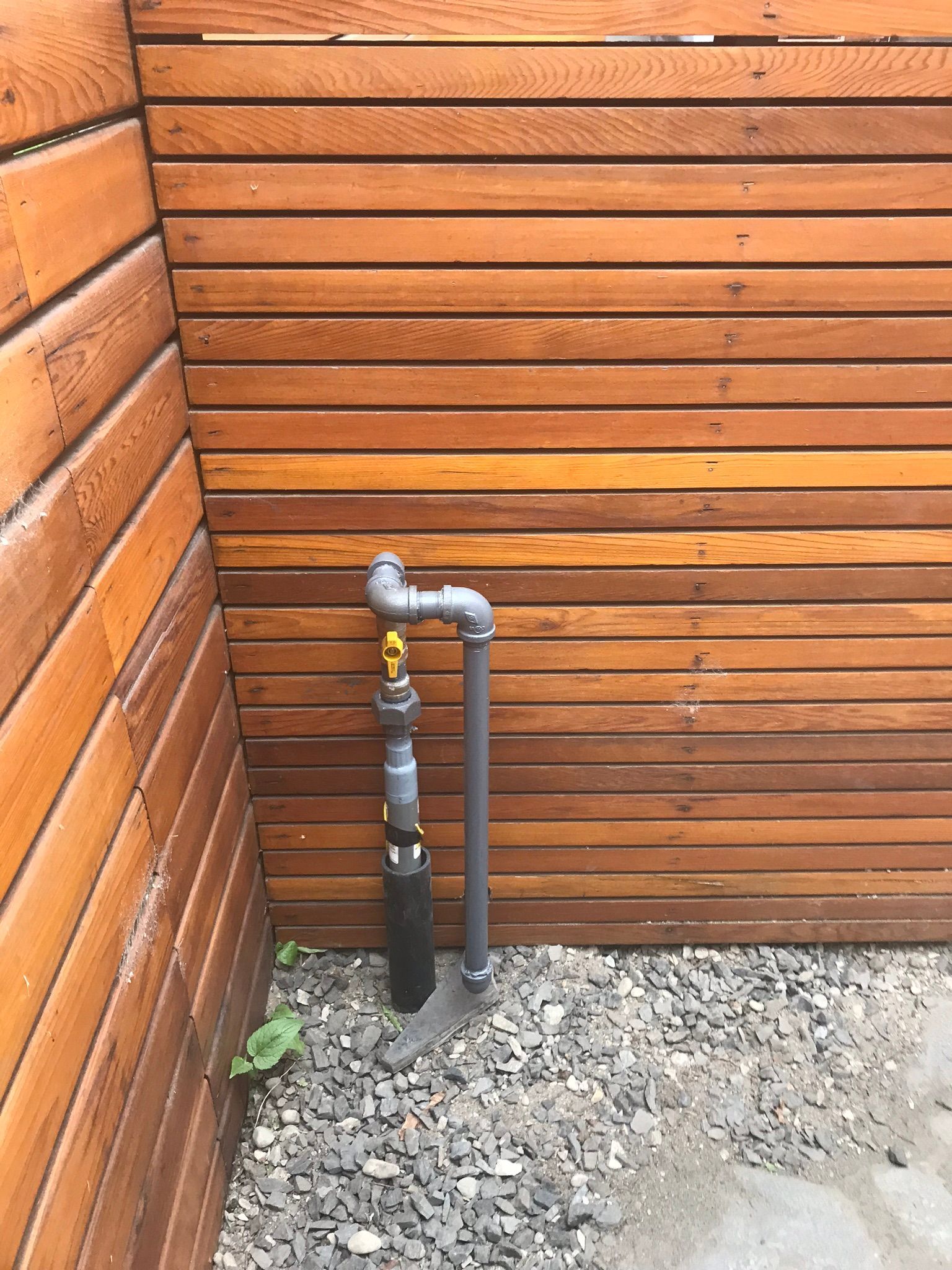 A pipe is attached to a wooden wall next to a gravel area.