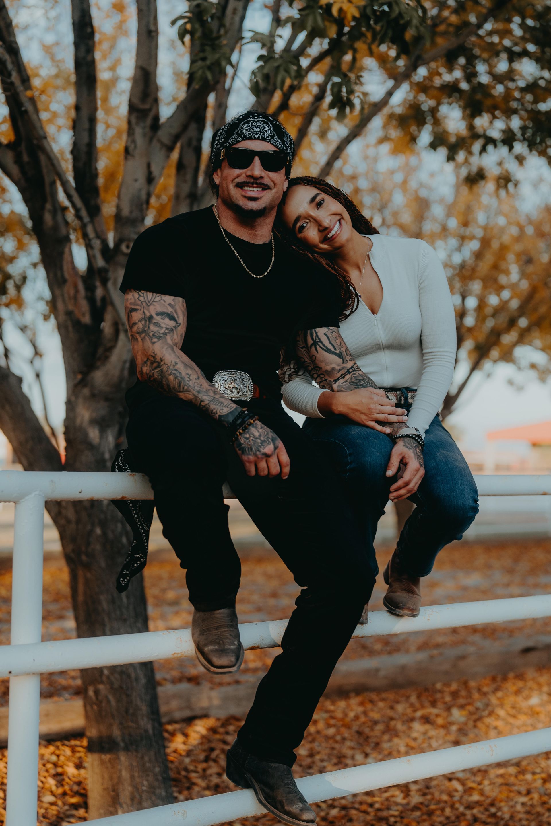 Couple sitting on a fence outdoors. Man with tattoos and a bandana, woman resting on his shoulder. Autumn colors.