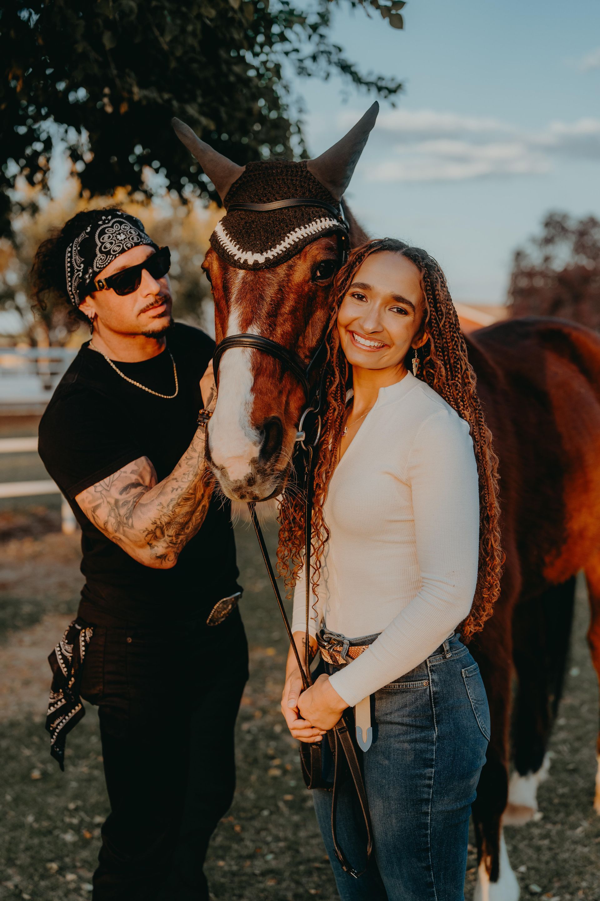Couple with a horse: man in black, woman in white. Horse wears decorated headgear. Outdoors, sunny day.