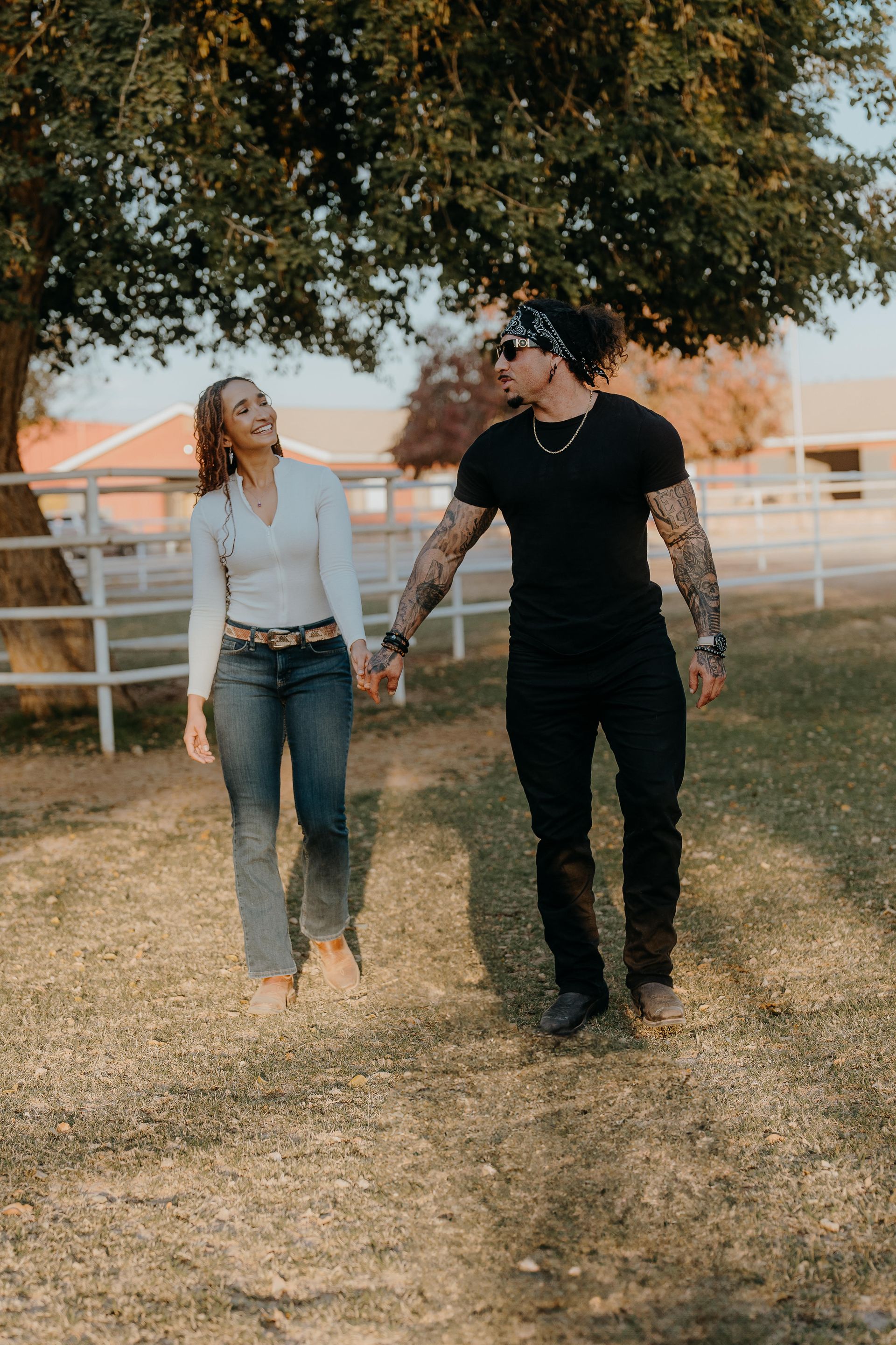 Couple holding hands, walking in a grassy field. Woman smiles, wearing jeans. Man has tattoos. Setting has trees, fence.