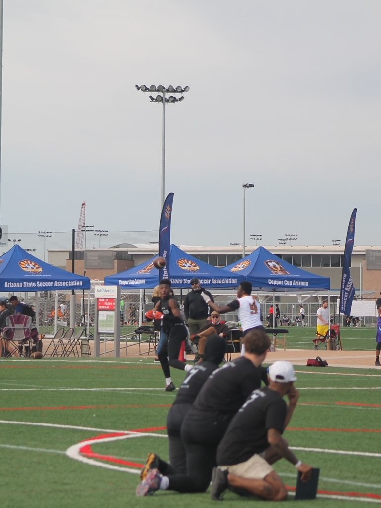 Football game on a green field with players, staff, tents, and sky.