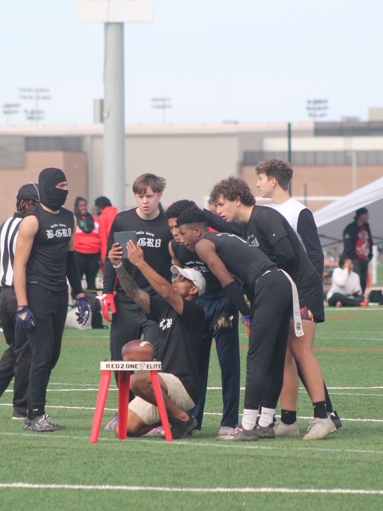 Group of young men huddle around a football on a football field. They are wearing athletic gear and looking at the ball.