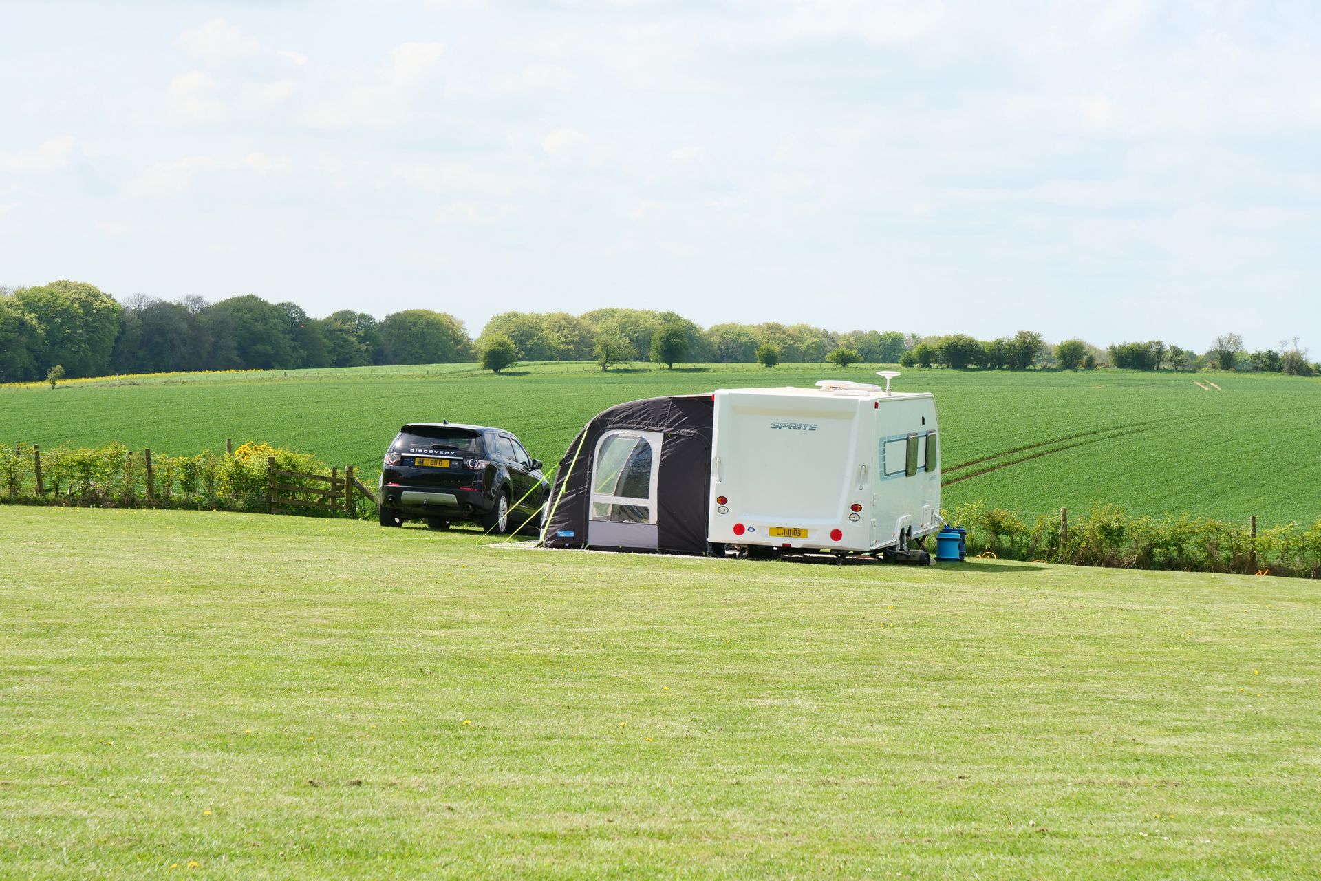 Black SUV and white camper parked on a grassy field beside green farmland under a bright sky