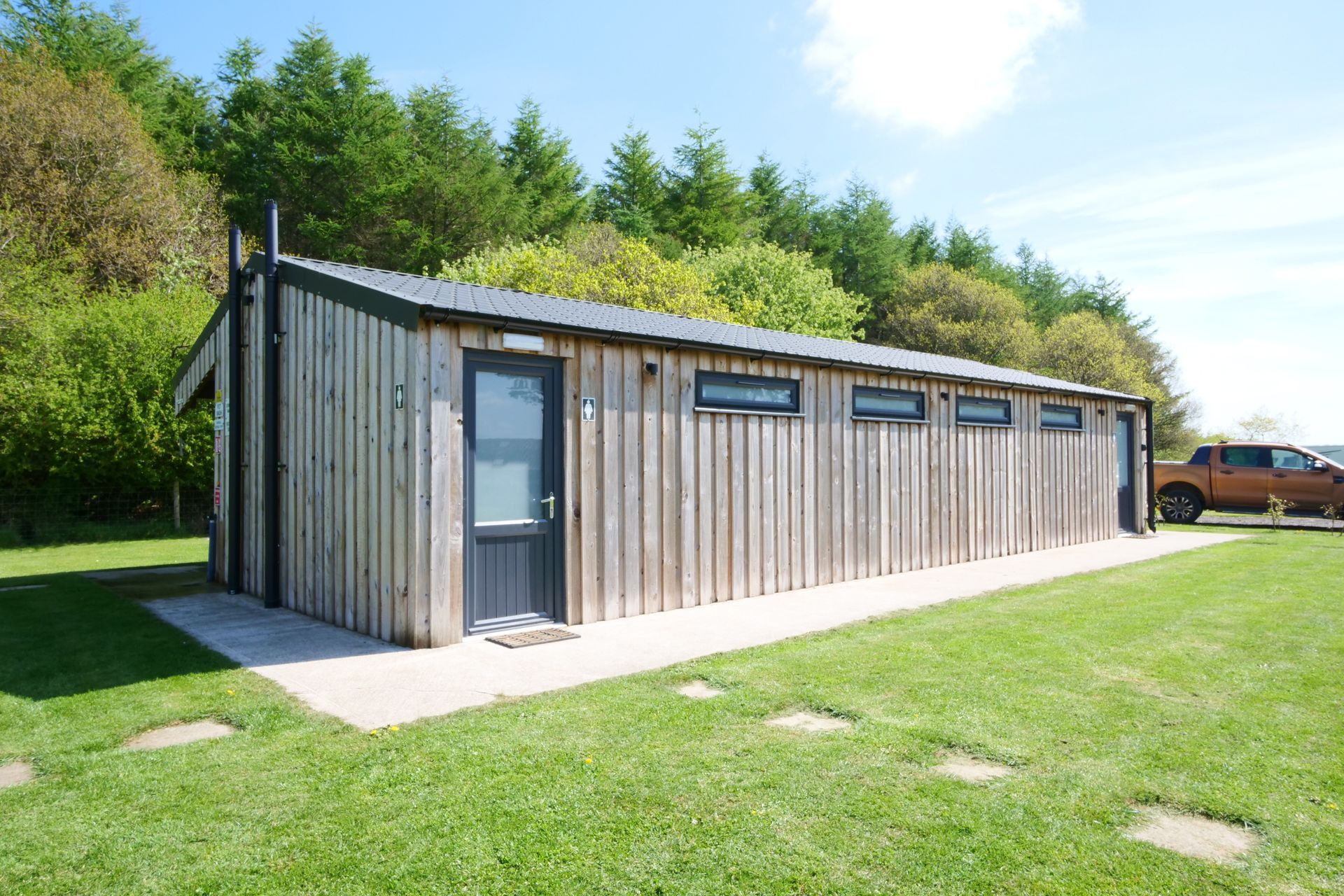 Small gray wooden shower block with a door and windows in a grassy park setting