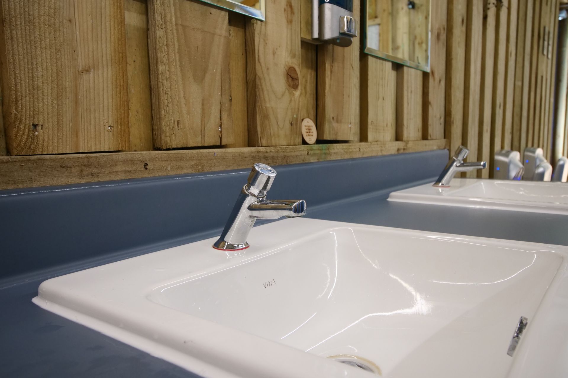 Two white bathroom sinks with chrome taps against blue counters and a wooden wall
