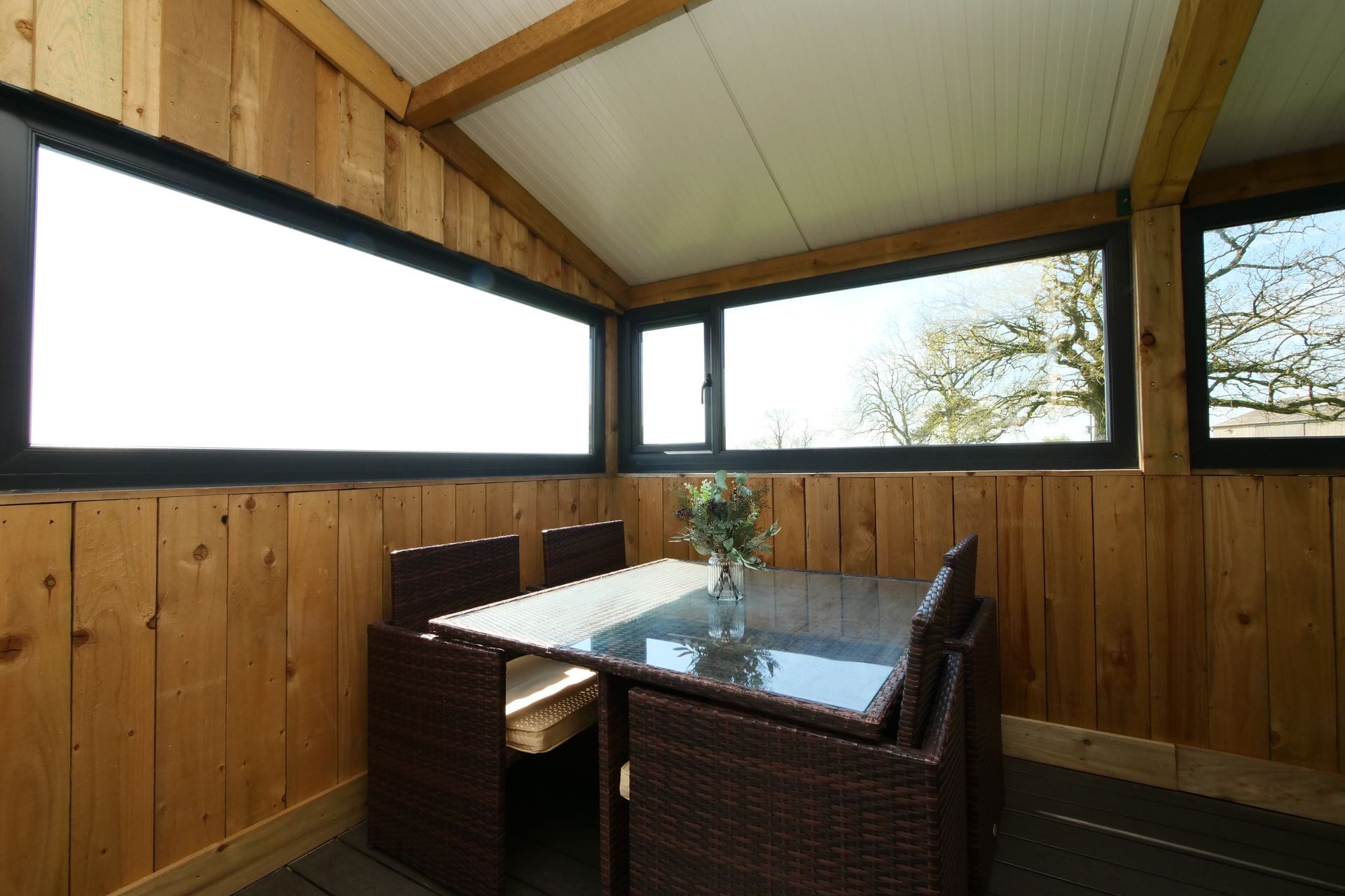 Pod with wicker dining set, glass-top table, and wood-paneled walls overlooking trees