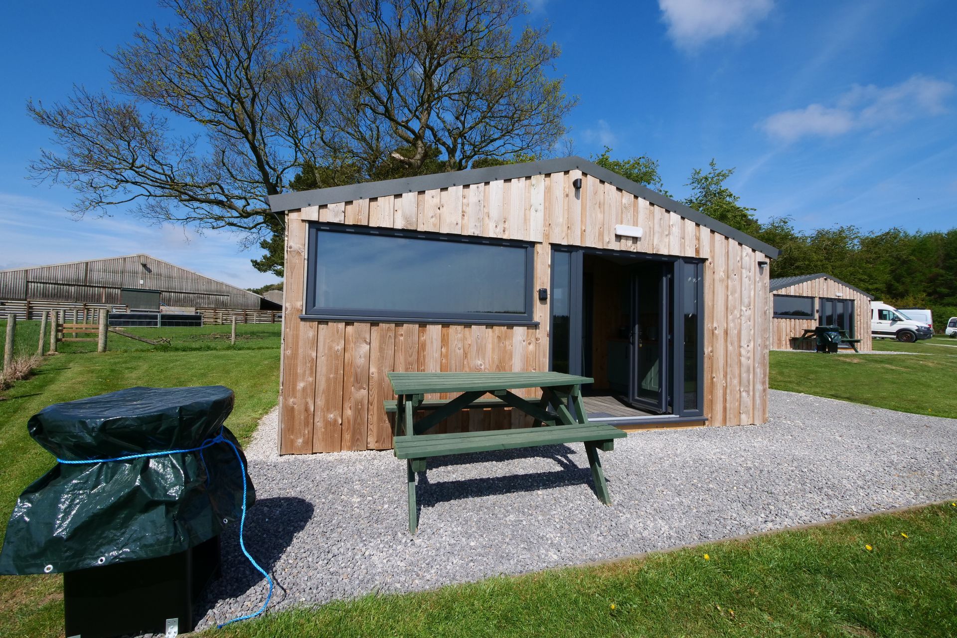 Small wooden cabin with picnic table on gravel, blue sky overhead, in a grassy field