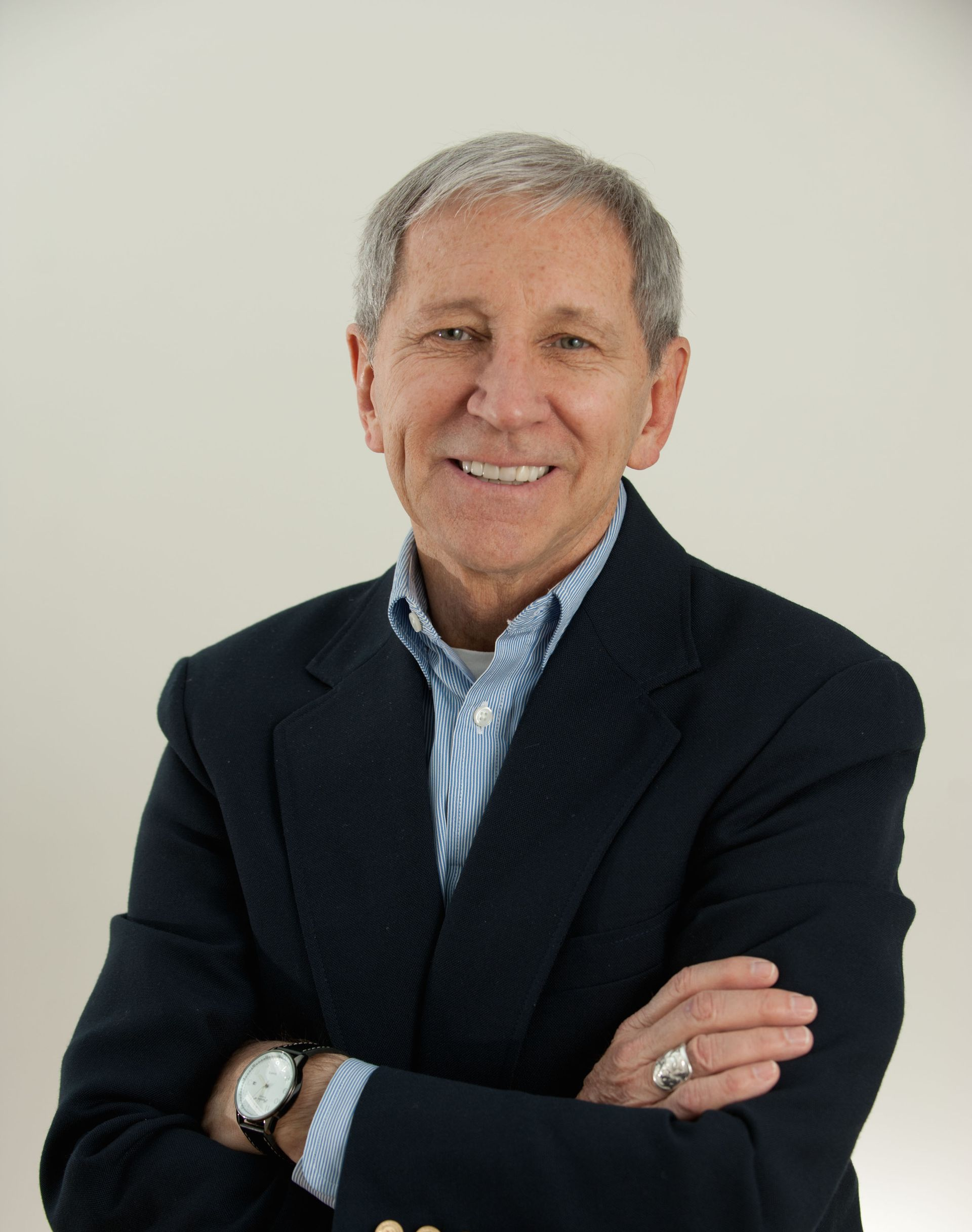 Man in a navy blazer and striped shirt, arms crossed, smiling against a light background.