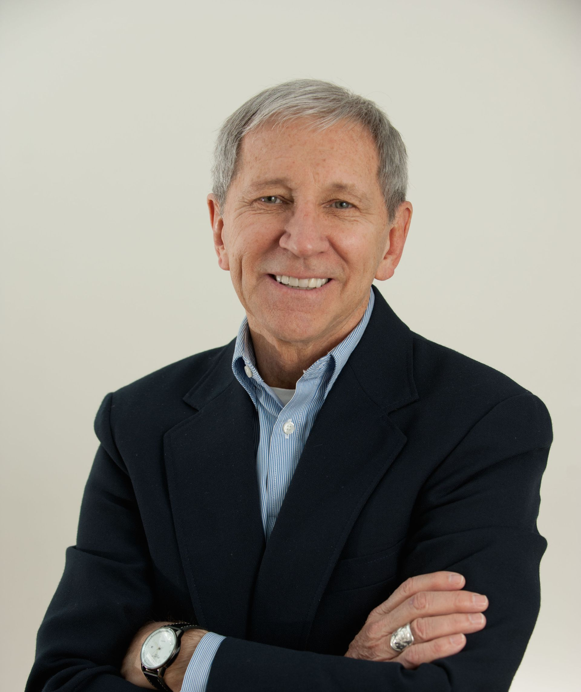 Man in a navy blazer smiles, arms crossed, wearing a watch. Pale background.