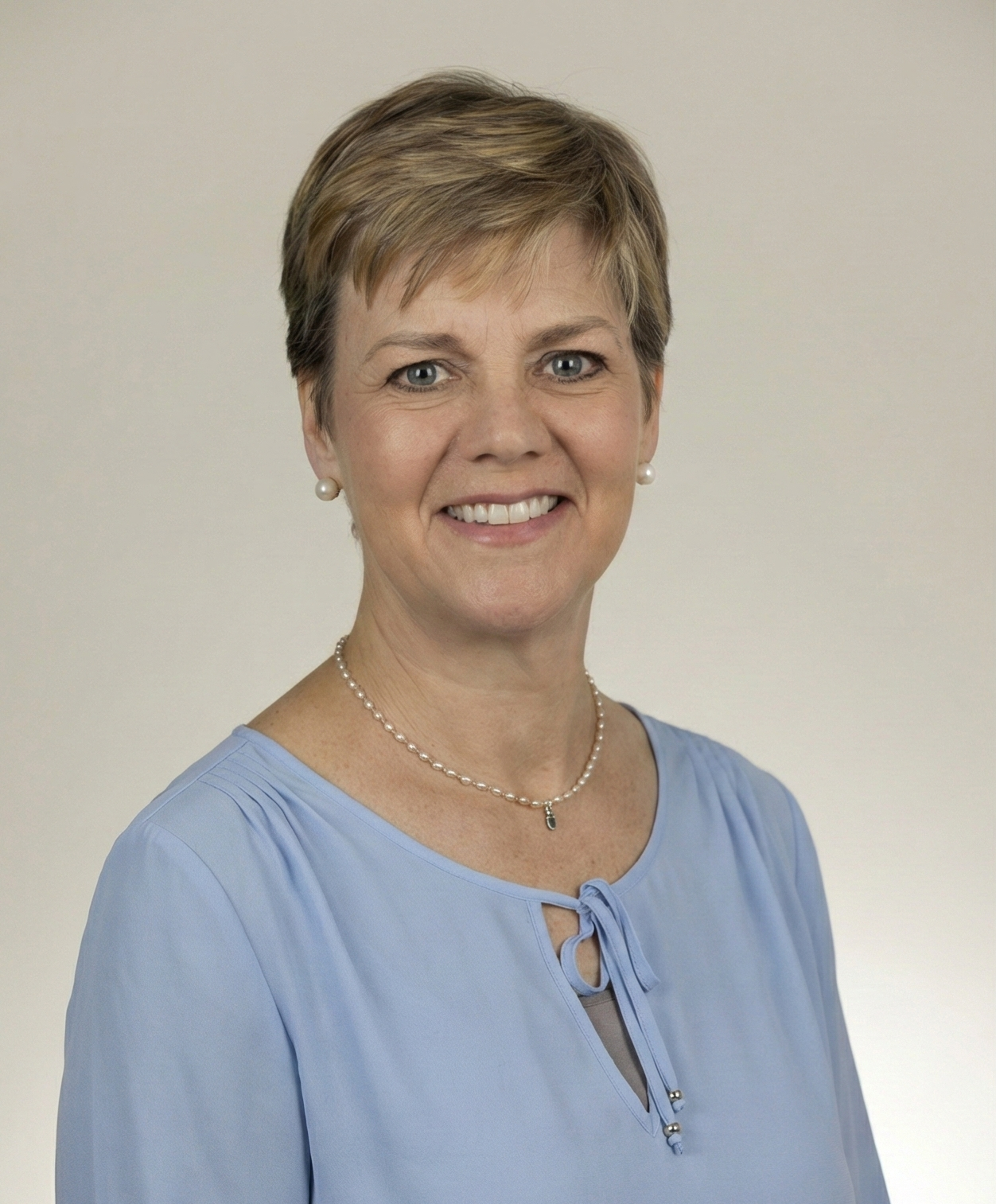 Woman with short blonde hair smiles, wearing a light blue top and a delicate necklace against a plain background.