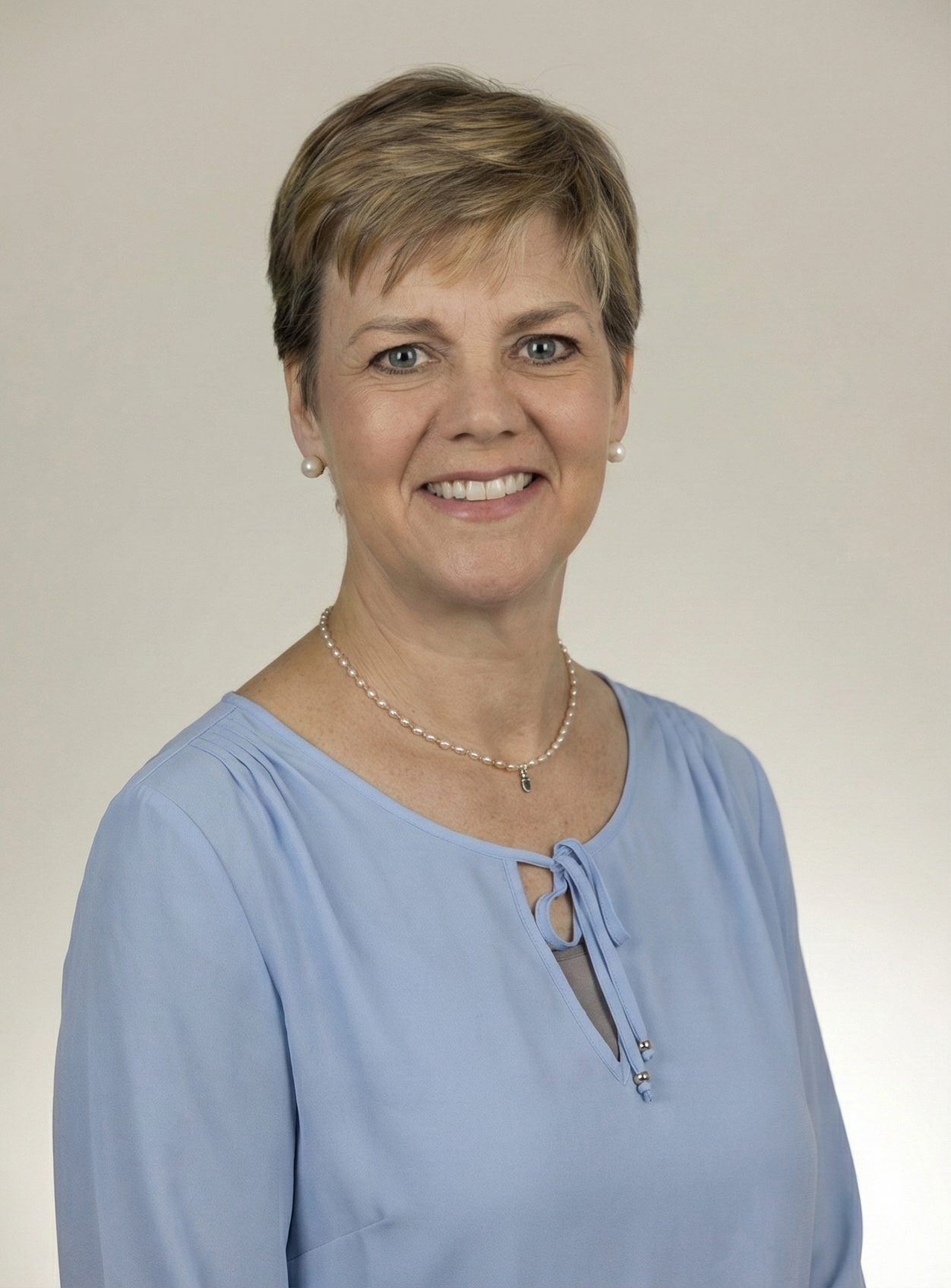 Woman with short blonde hair smiles, wearing a light blue top and pearl necklace.
