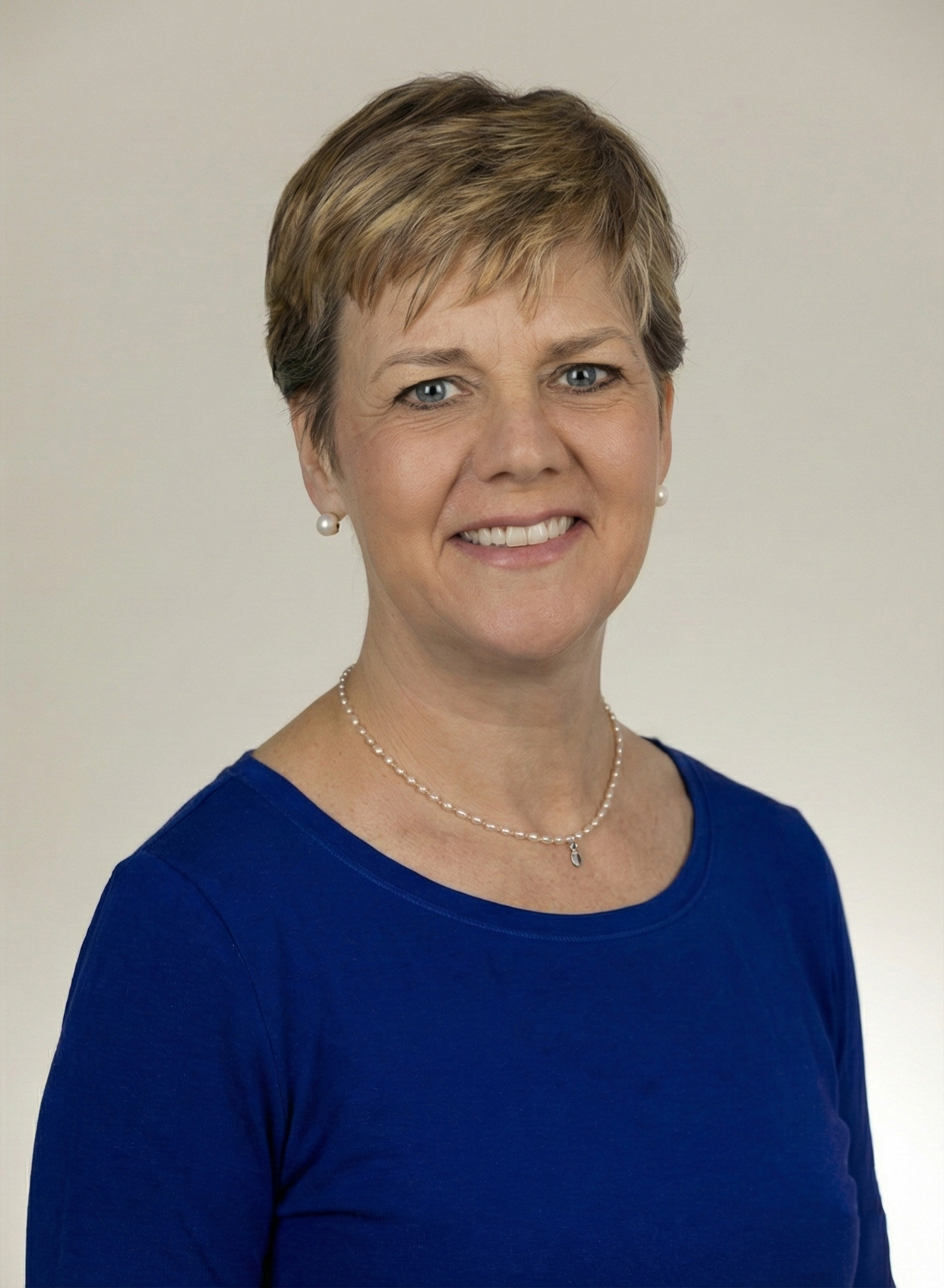 Woman with short blonde hair smiles, wearing a blue shirt and necklace, against a light gray background.