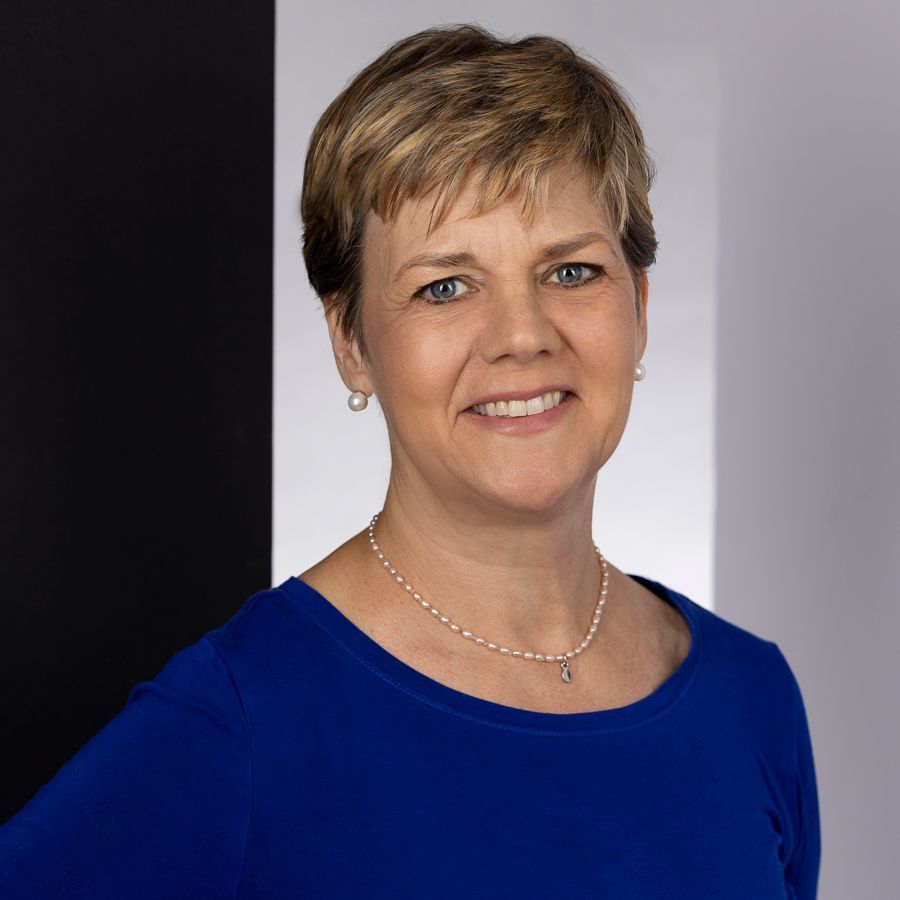 Woman with short blonde hair in a blue top smiles, wearing a pearl necklace; in front of a black, white, and gray background.