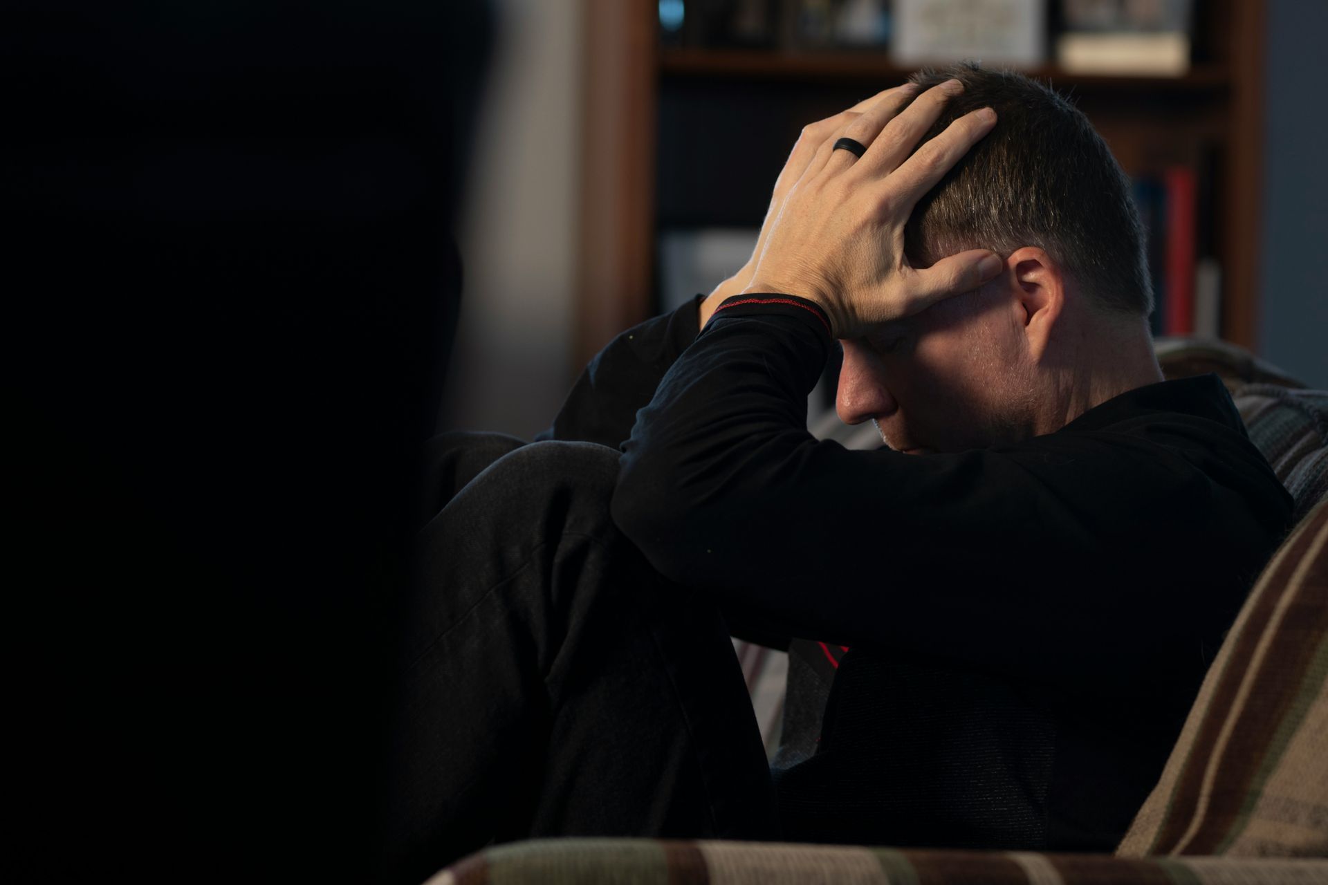 Man with hands on head, seated on a couch, appearing distressed; bookshelf in background.