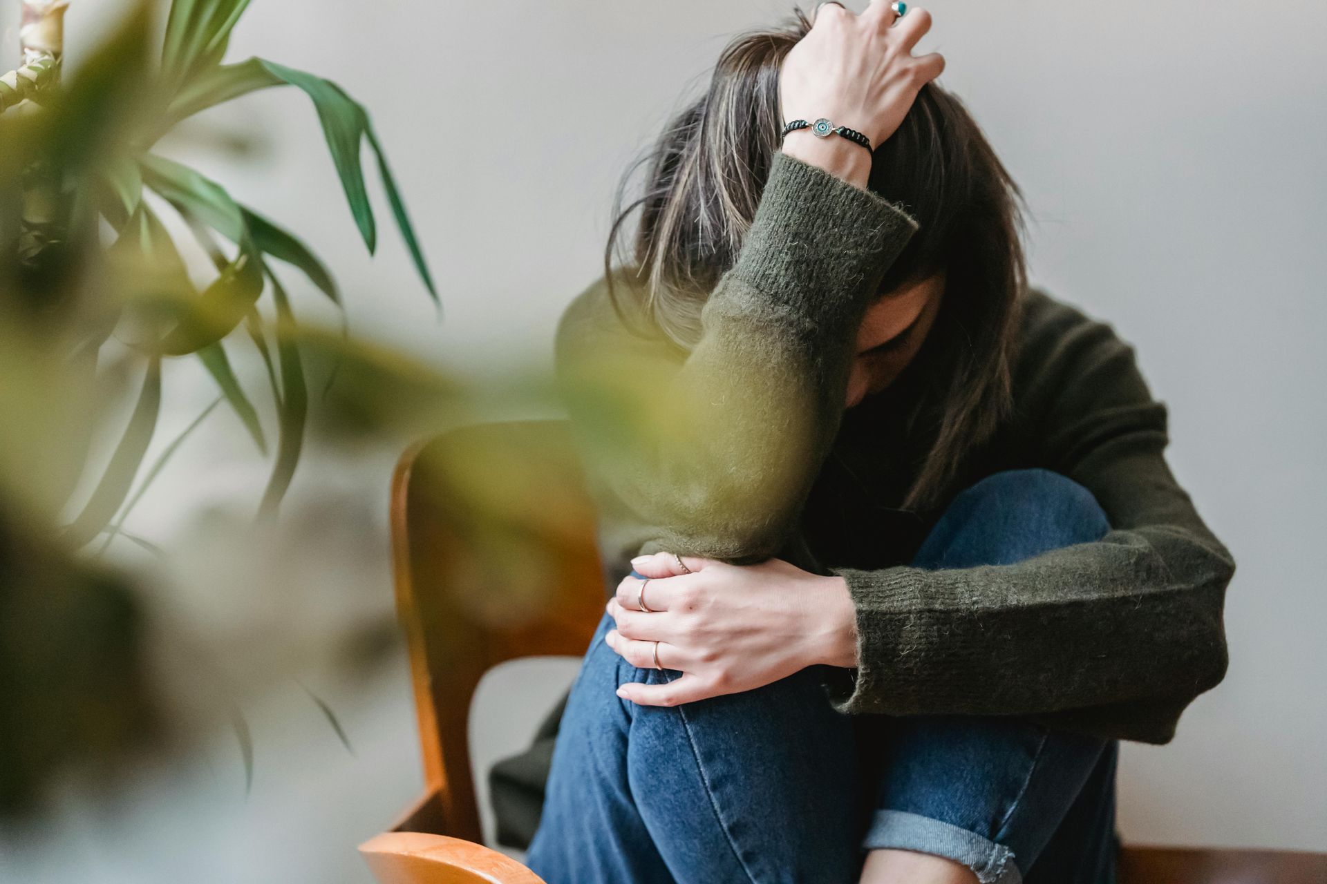 Woman hunched over, head in hands, sitting in a chair. Wearing jeans and a green sweater, indoors.
