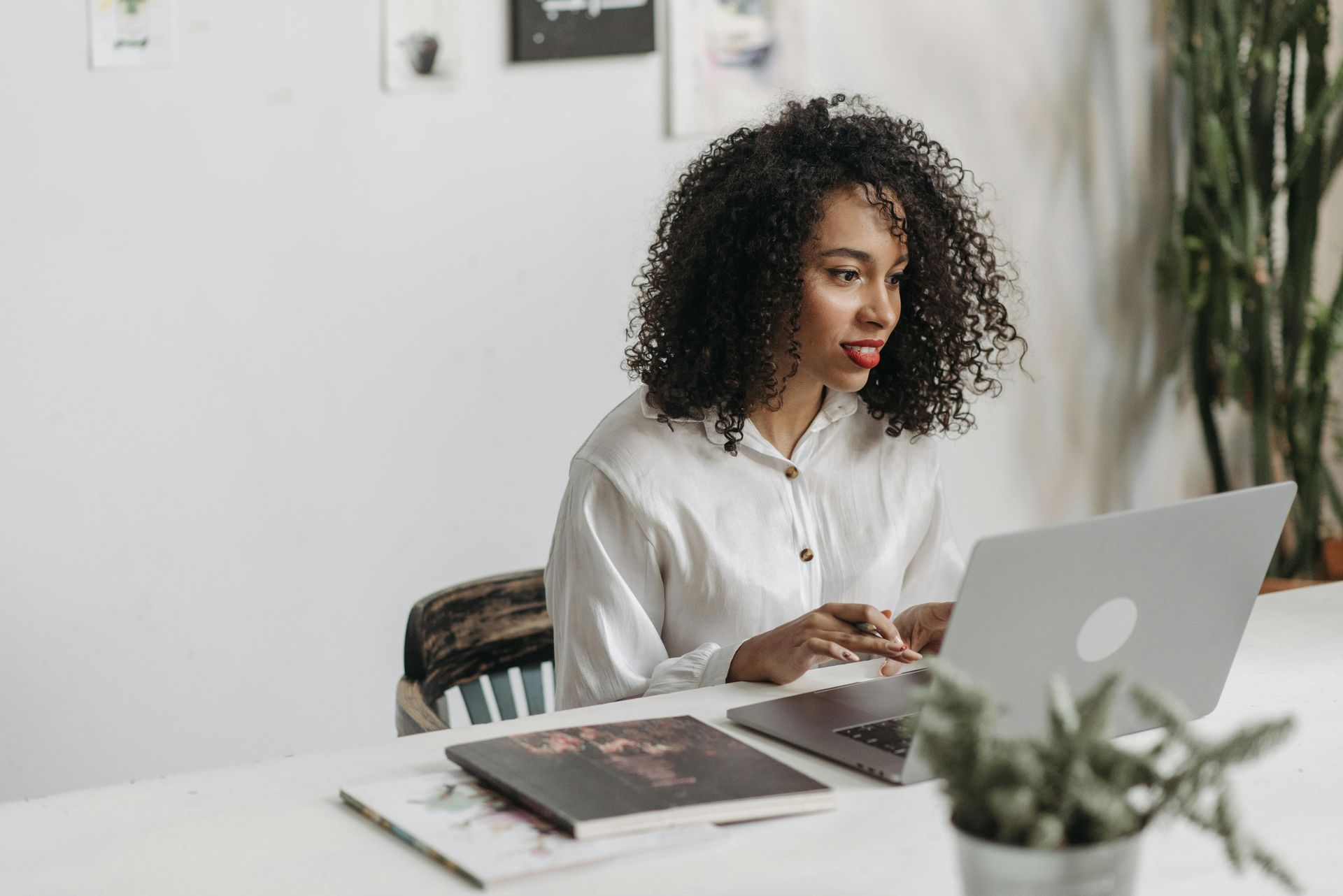 Woman with curly hair, in white shirt, working on laptop at desk, focused expression.
