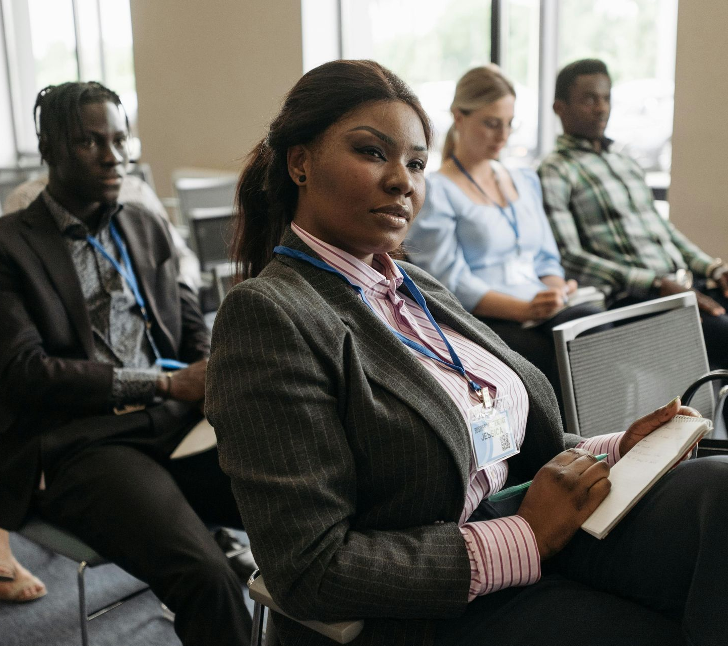 Attendees at a conference. Woman in blazer looks at camera, holding notebook. Others listen.