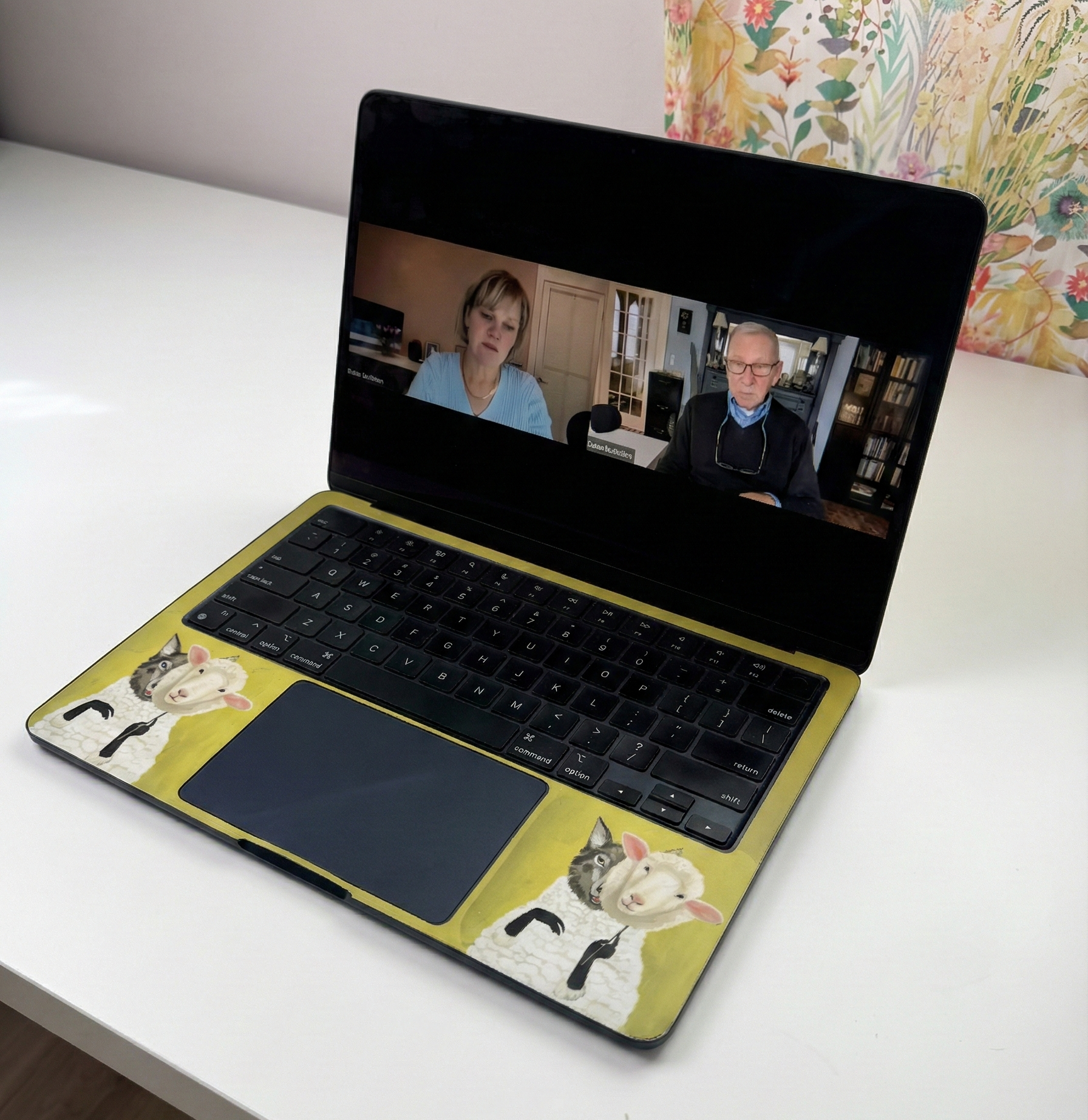 Laptop on a white table displaying a video call with two people. The laptop has cat stickers.