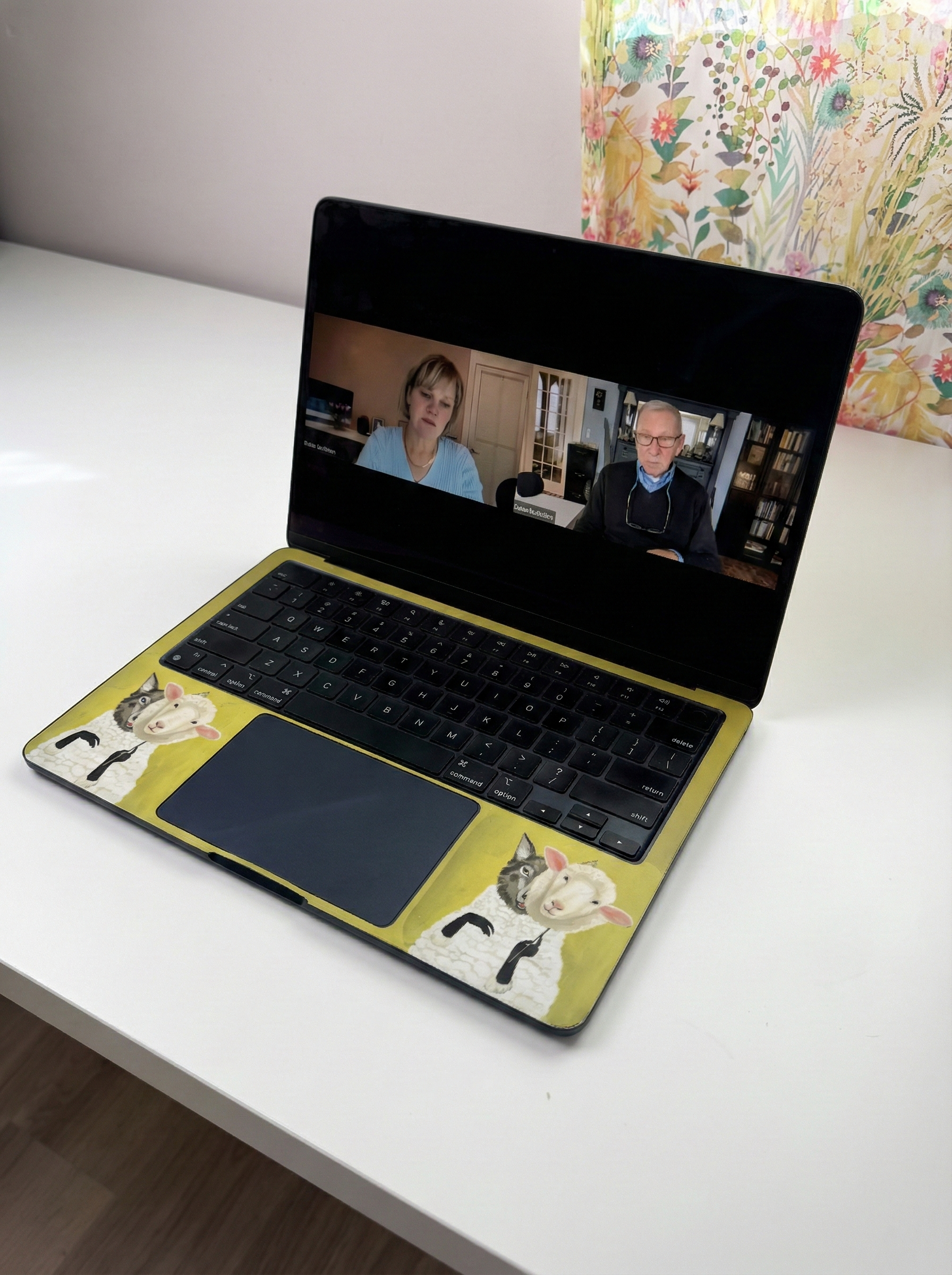 Laptop on a white desk displays a video call with two people. The laptop has cow stickers.