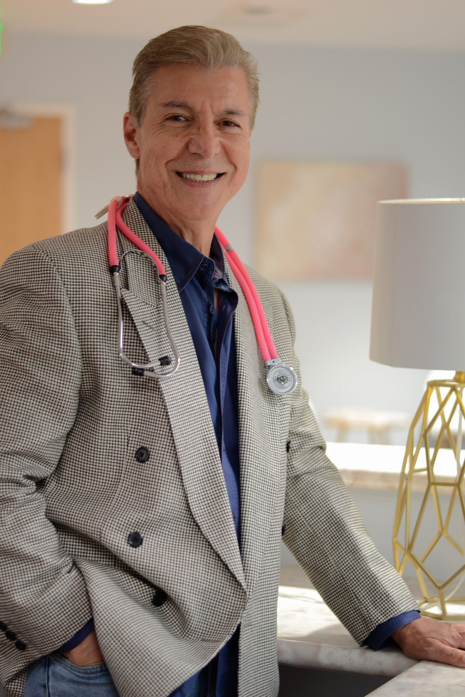 Man in patterned blazer with medals, smiling beside a lamp in a bright room