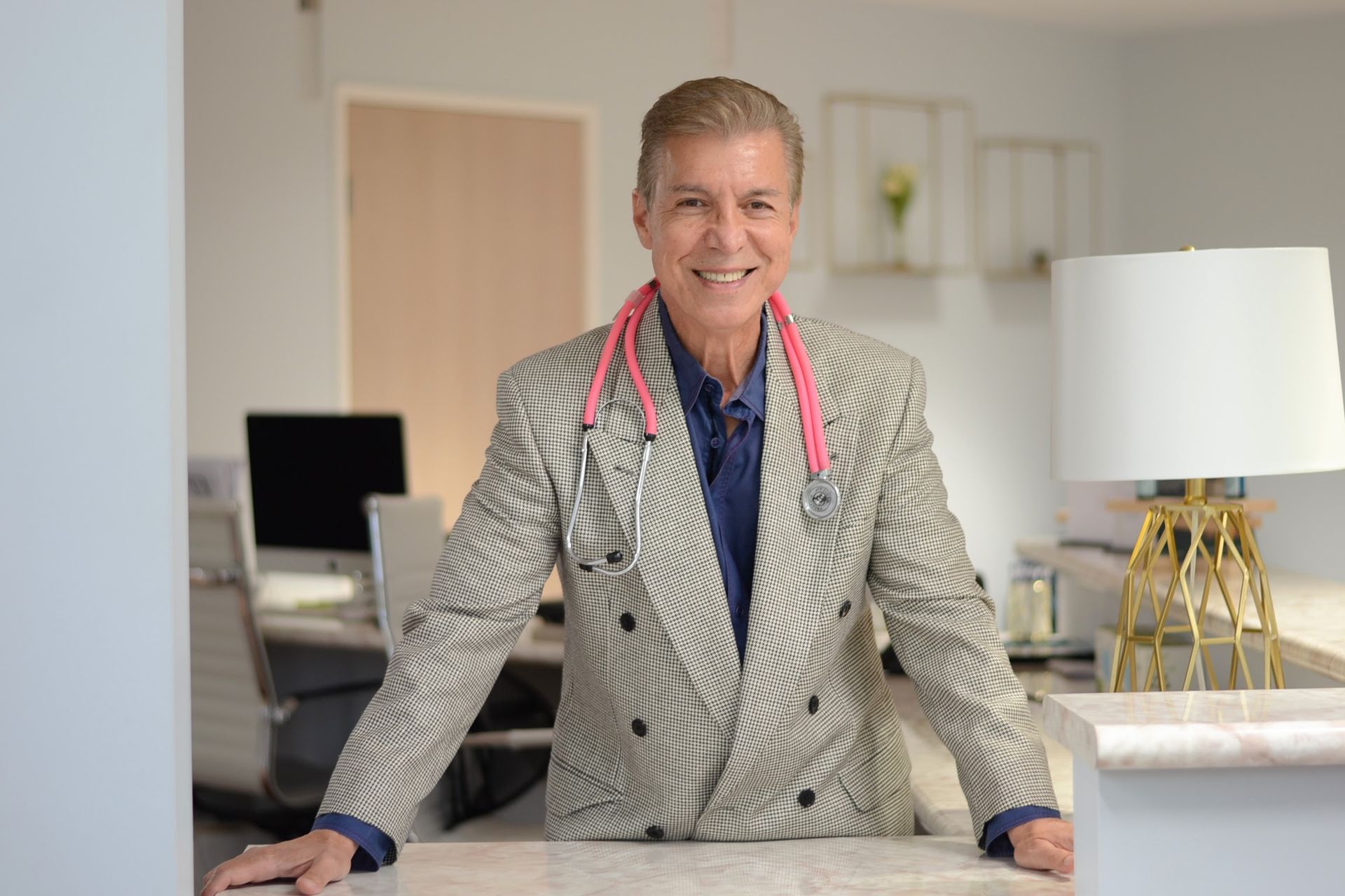 Smiling person in a patterned blazer standing in a bright modern room with a lamp and desk behind.