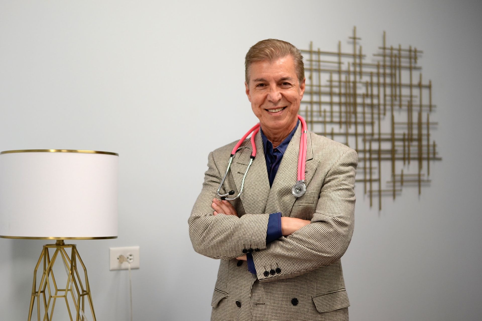 Smiling person in a patterned suit stands with arms crossed beside a lamp and wall art.