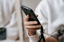 Person holding a black phone with a wrist strap, seated indoors against a light background