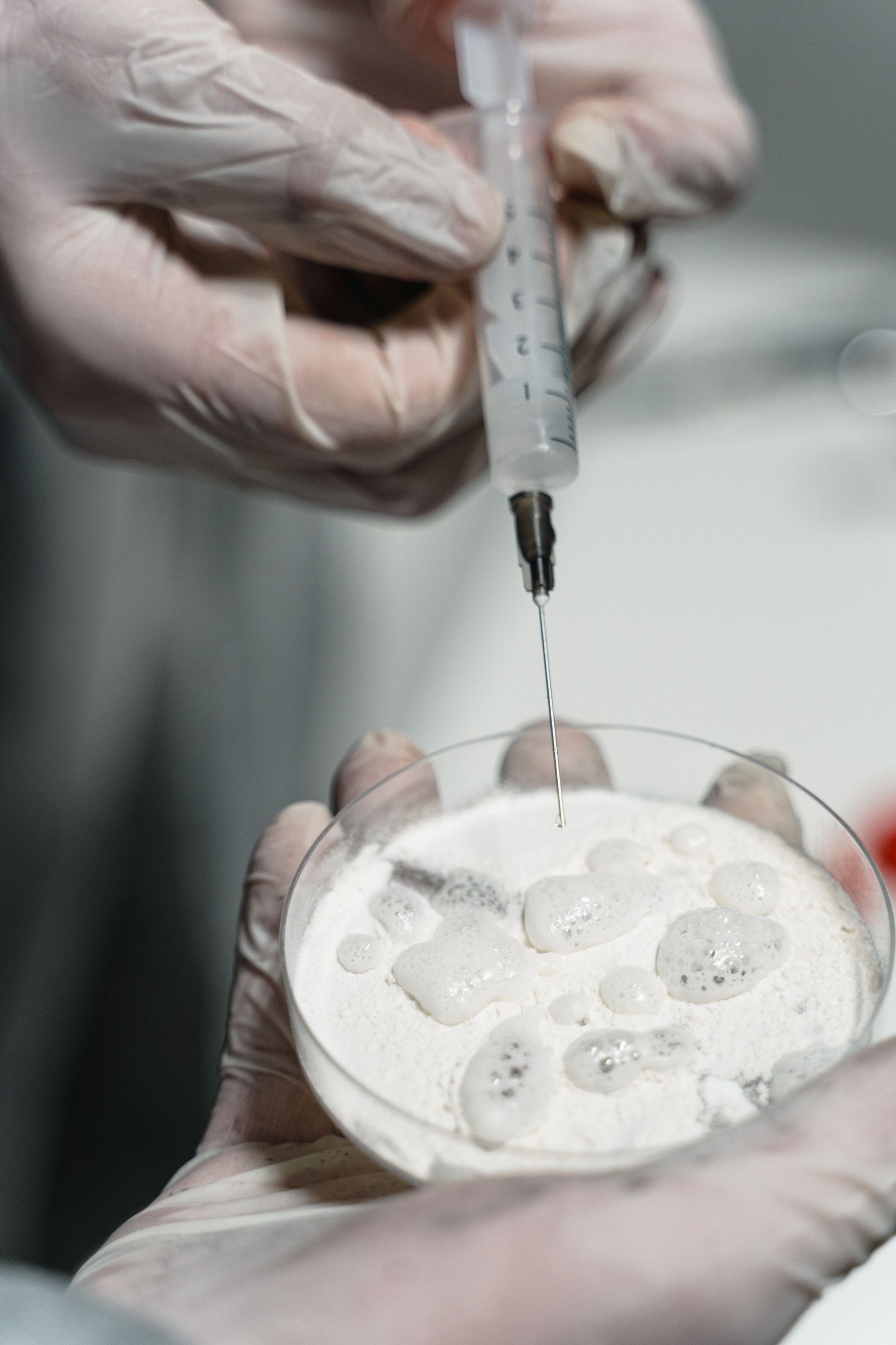 Gloved hands use a syringe to dispense liquid into a petri dish of samples in a lab