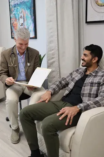 Two men chatting in a bright office lounge, one holding an open folder while seated in a cream chair.