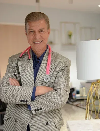 Smiling doctor with stethoscope and pink scarf, standing with arms crossed in a bright office.