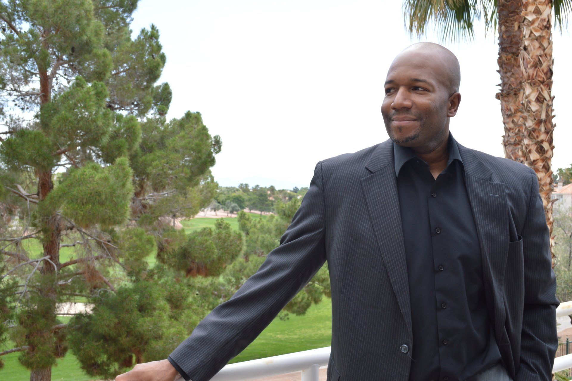 A man in a suit and black shirt is standing on a balcony with trees in the background.