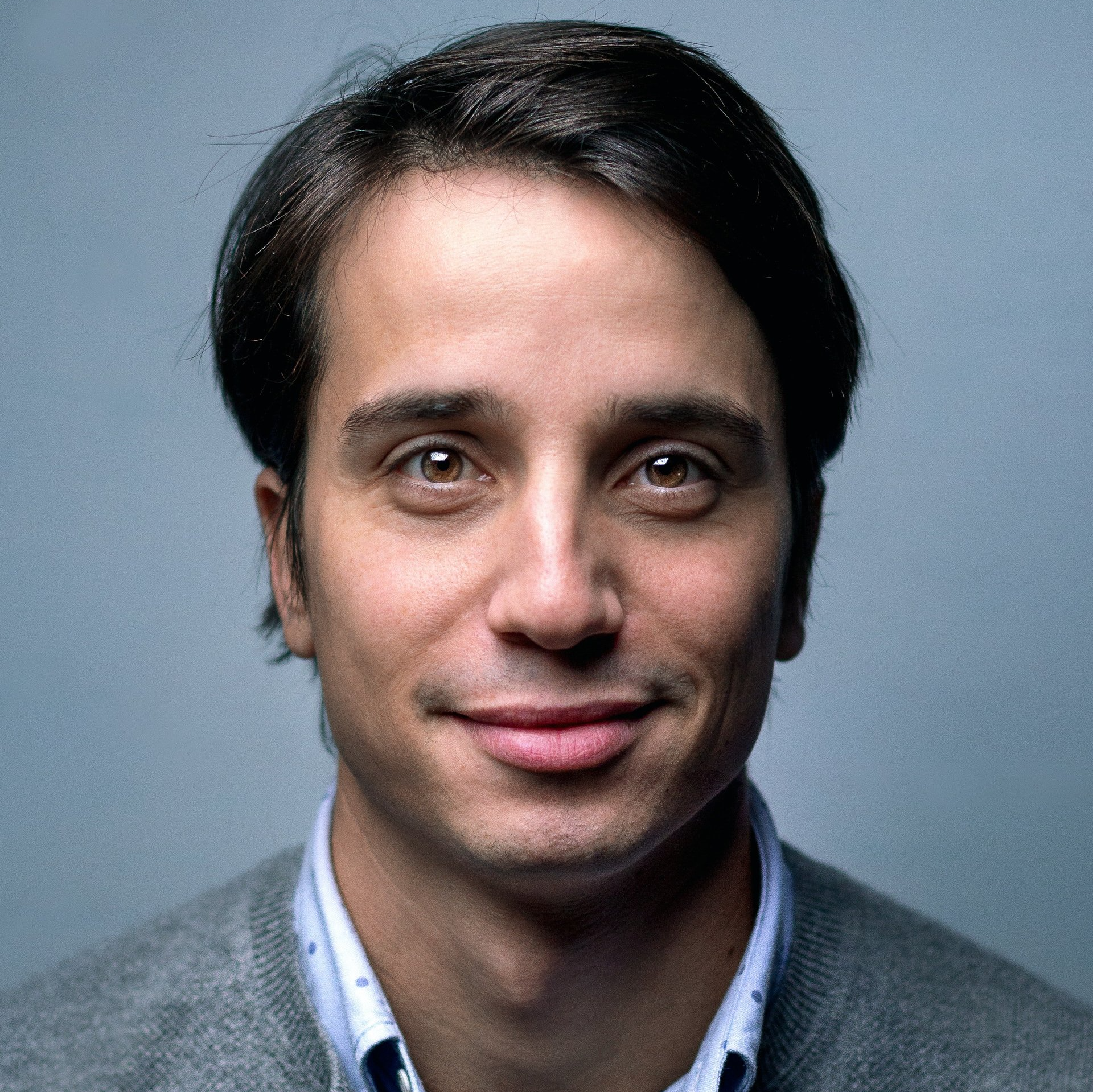 A man wearing a grey sweater and a white shirt is smiling for the camera.