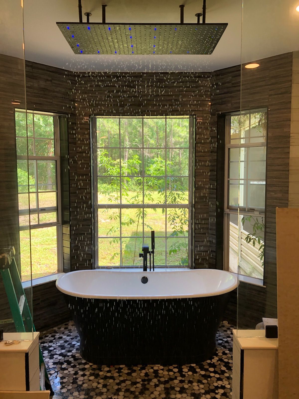 A modern bathroom with a freestanding black textured bathtub centered in front of three windows under a large rain shower.