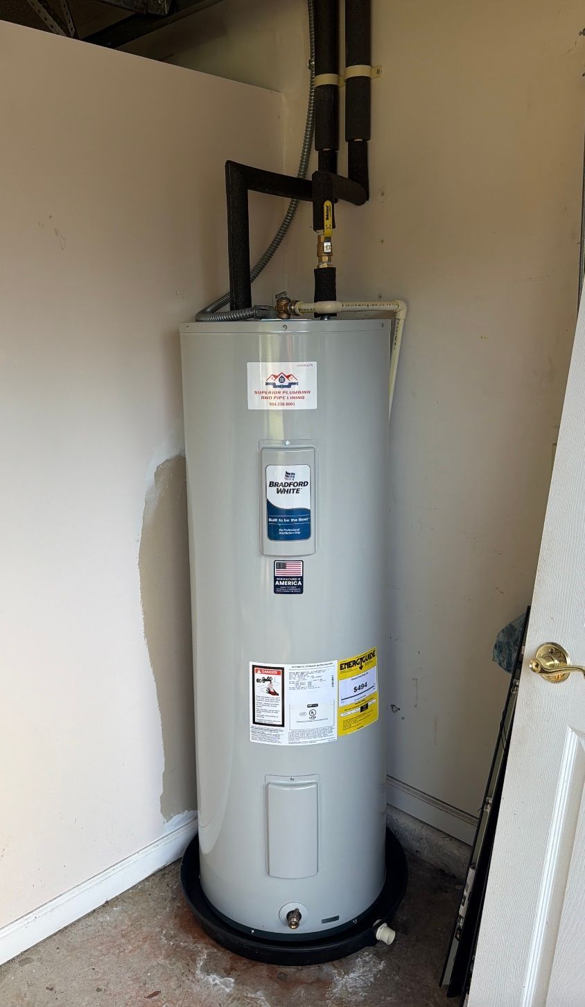 Tall gray water heater in a utility closet with exposed black pipes and beige walls.