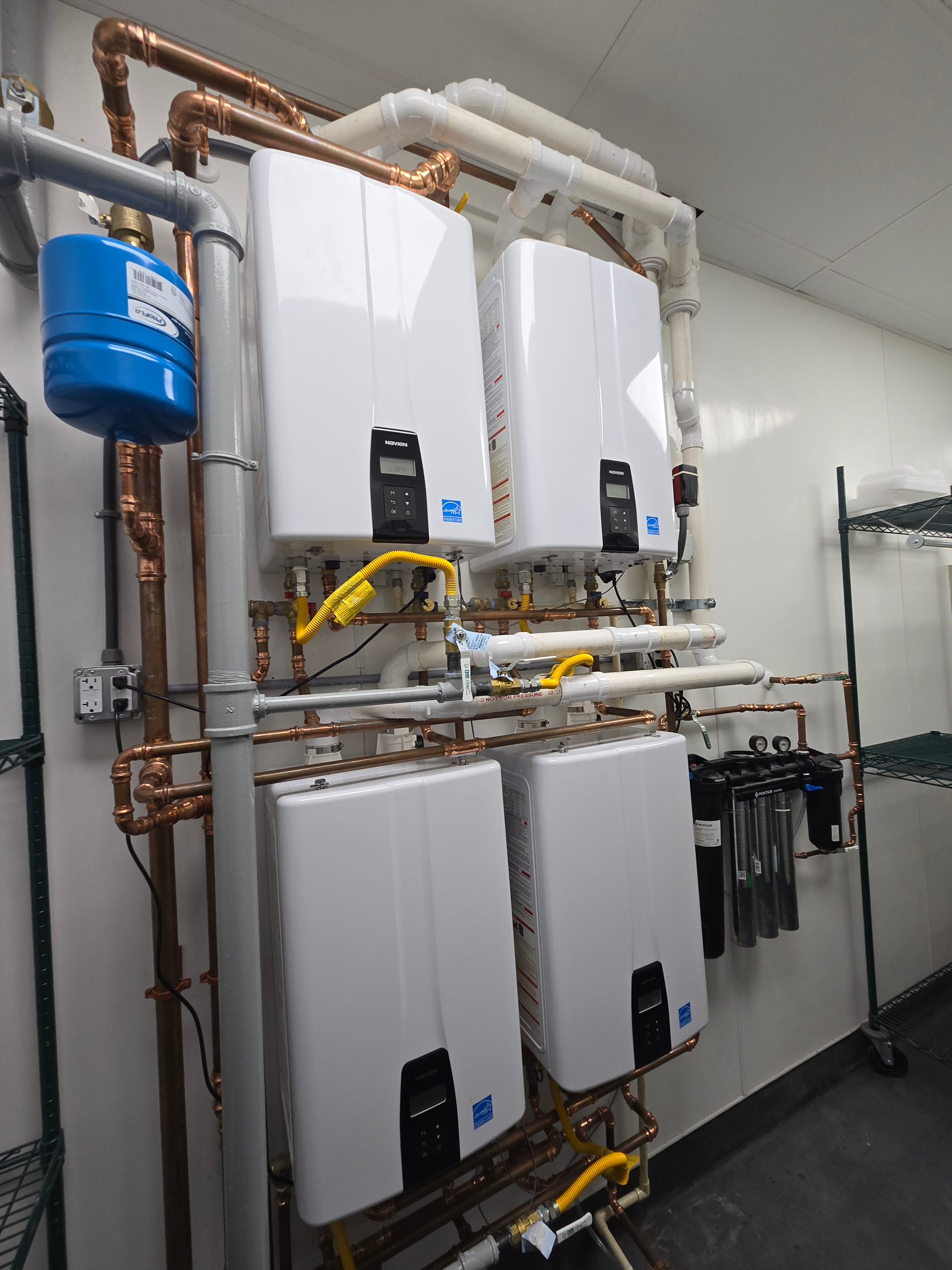 A wall-mounted system of four white commercial tankless water heaters connected by copper and white PVC piping.