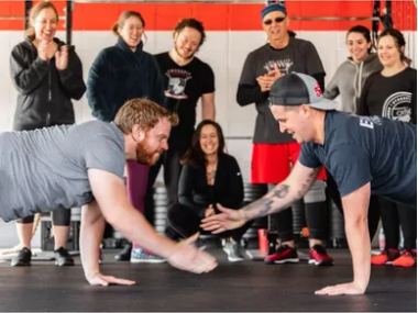 A group of people are standing around a man doing push ups in a gym.