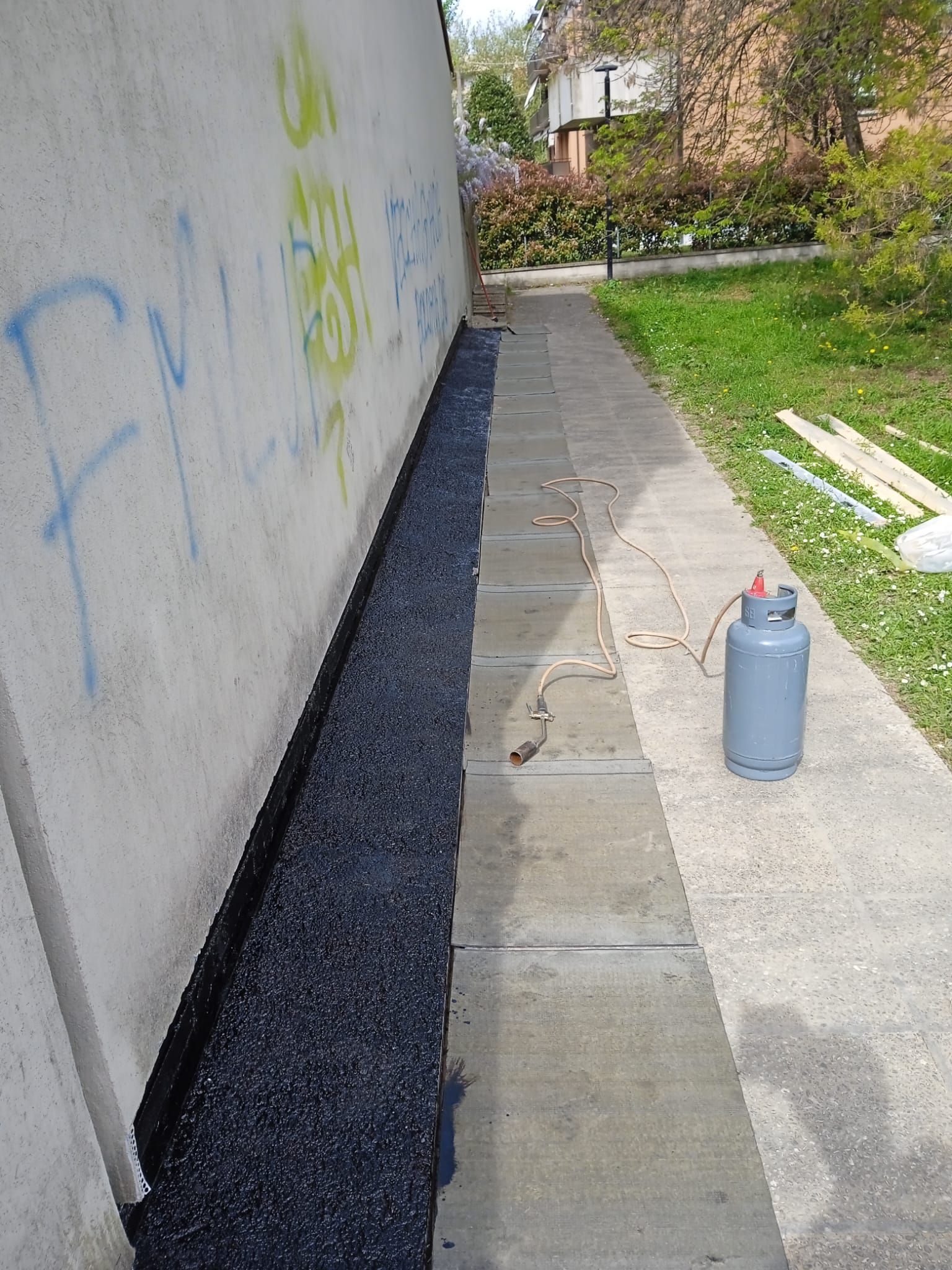 Una bombola di propano è appoggiata su un marciapiede, accanto a un muro decorato con graffiti.