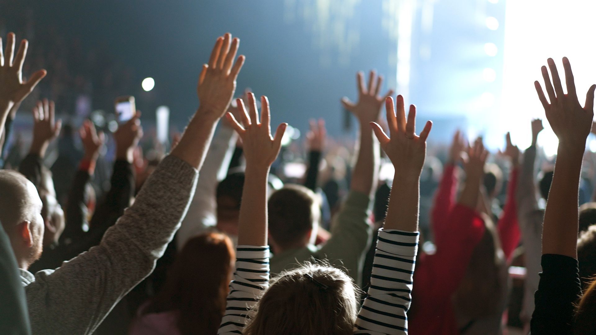 Crowd with raised hands at a concert, bright stage lights in the background