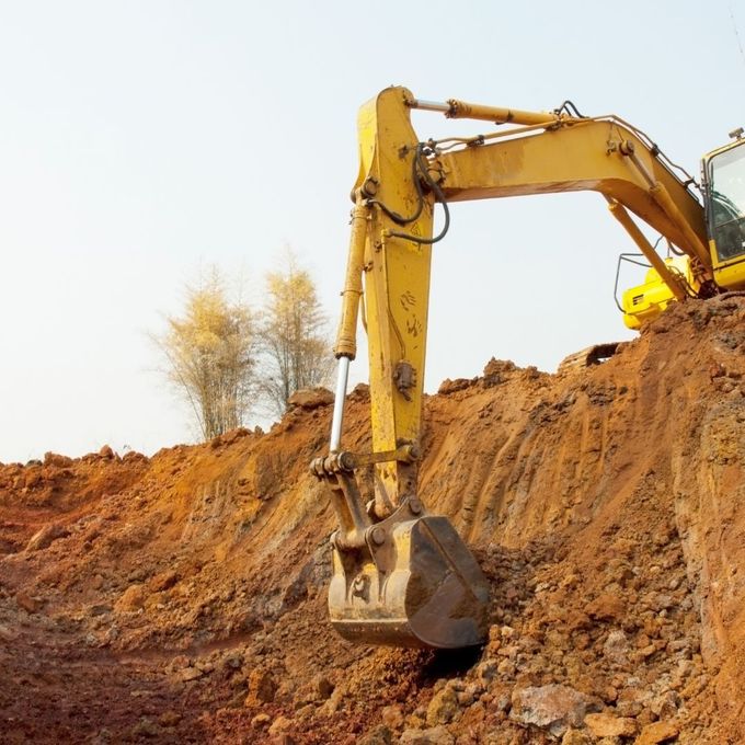 Yellow excavator digging into a mound of reddish-brown dirt against a light sky.