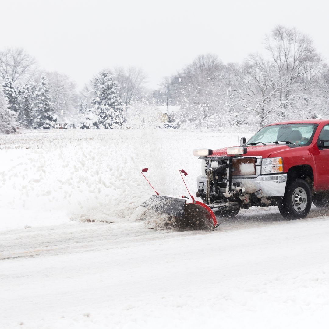 Red truck plowing snow from a road on a snowy day; trees in the background.