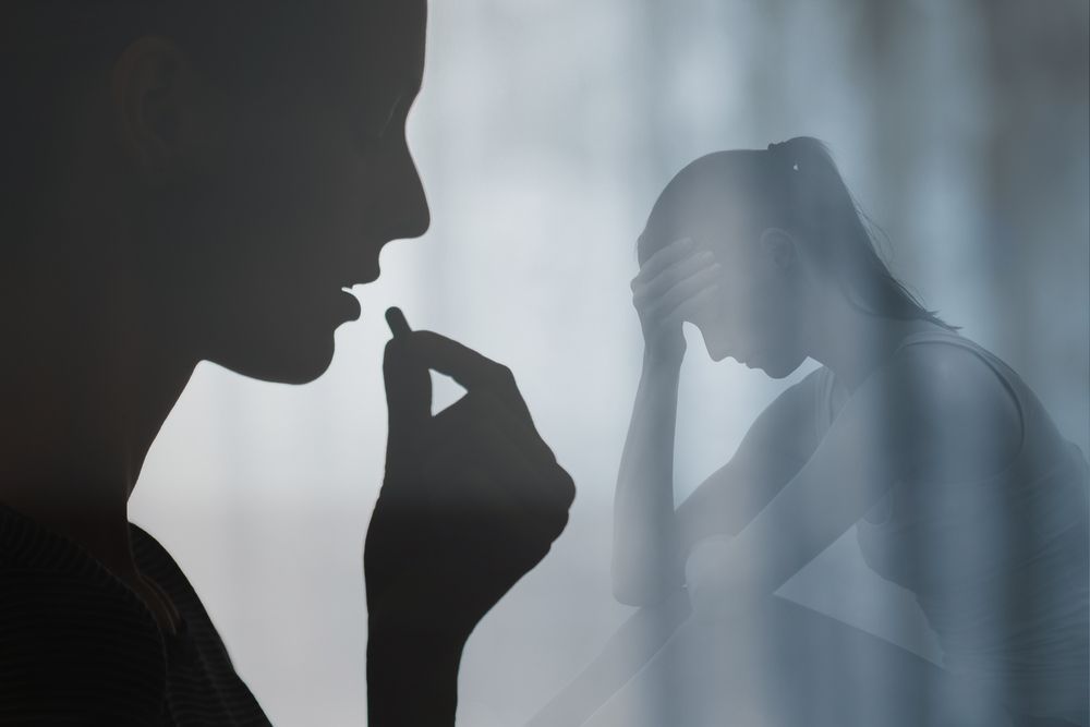 Silhouette of a person taking a pill while another person sits nearby, head in their hands in distress.