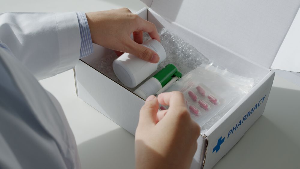 A person in a white lab coat packing medical supplies, including a bottle and blister-packed pills, into a branded box.