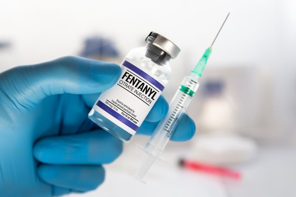 A hand wearing a blue medical glove holds a vial of fentanyl injection next to a prepared syringe.