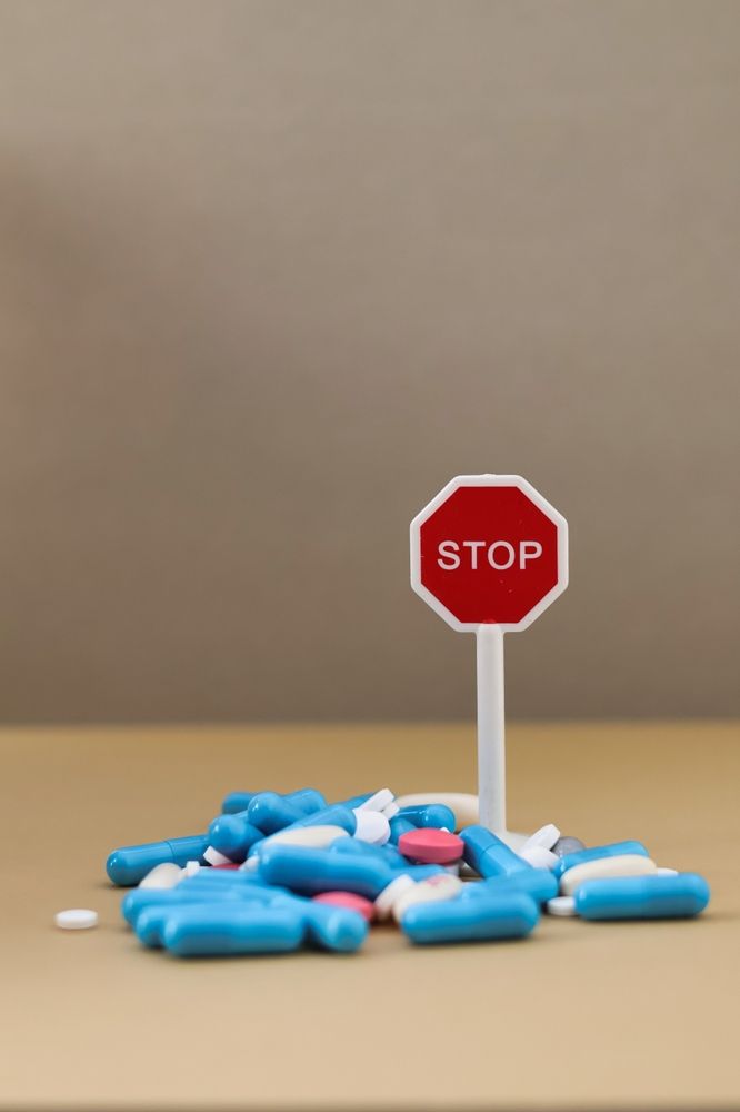 A pile of various blue and white medical capsules surrounding a small red and white stop sign on a neutral background.