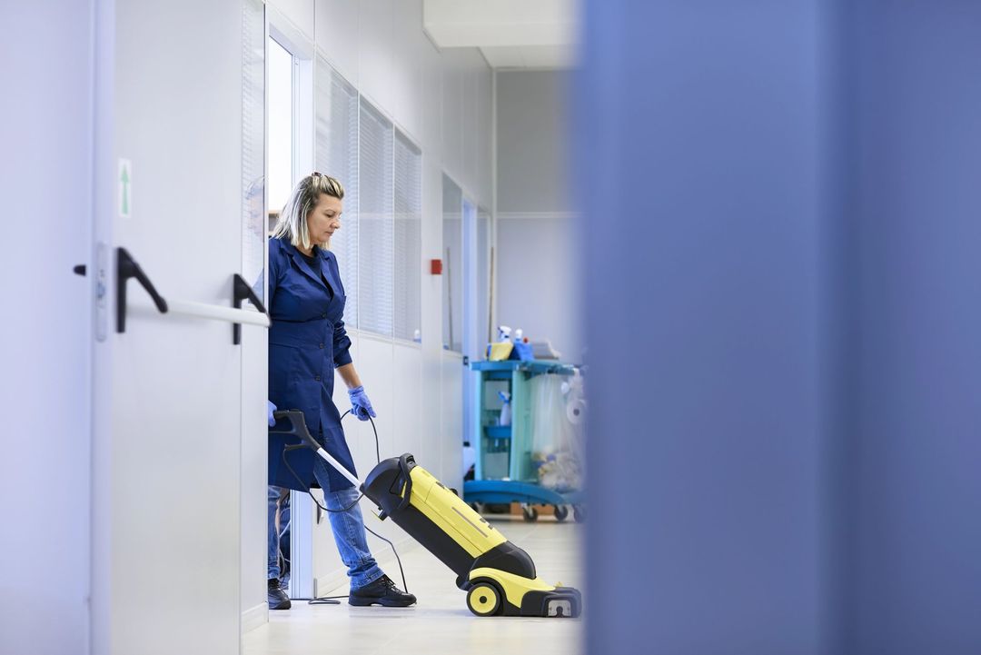 Addetto alle pulizie in uniforme blu che utilizza una macchina lavapavimenti gialla in un corridoio luminoso.