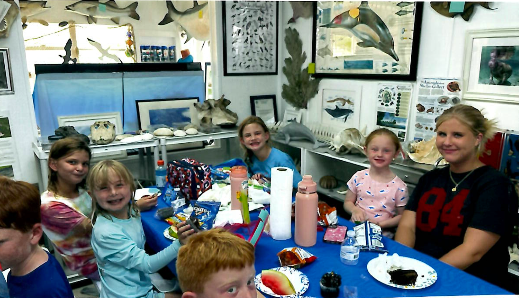 People in shallow water use a net. Three young women hold the net open near a pontoon boat on a cloudy day.