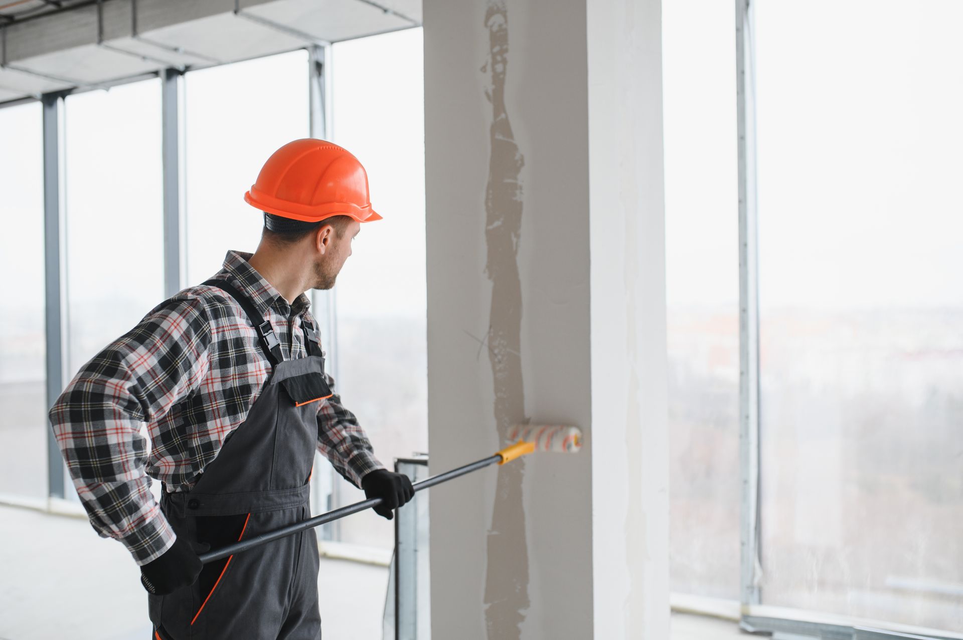 Construction worker painting a wall with a roller; wearing a hard hat and overalls near a large window.
