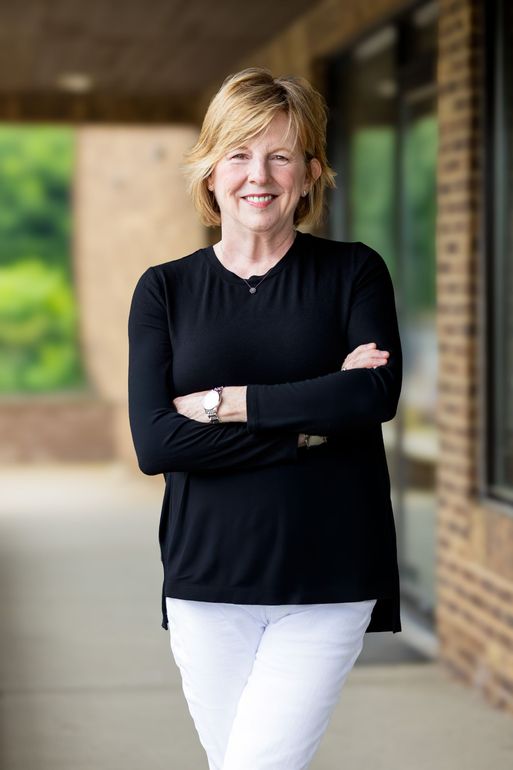 Woman with crossed arms, smiling, in black shirt and white pants, standing outside a building.