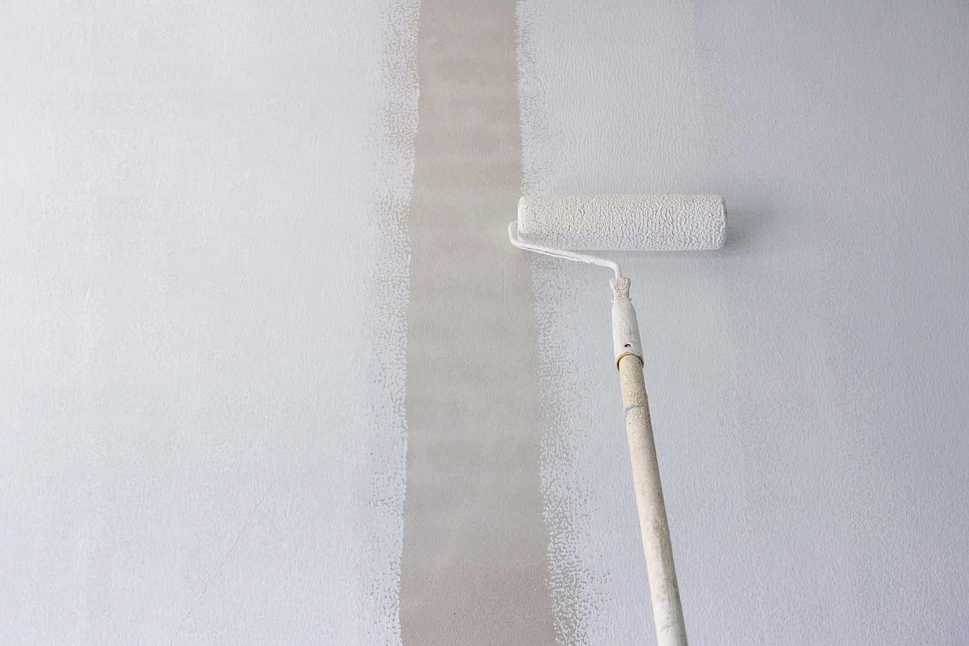 An individual using a long handle roller brush to apply a coat of primer white paint onto a cement wall background.