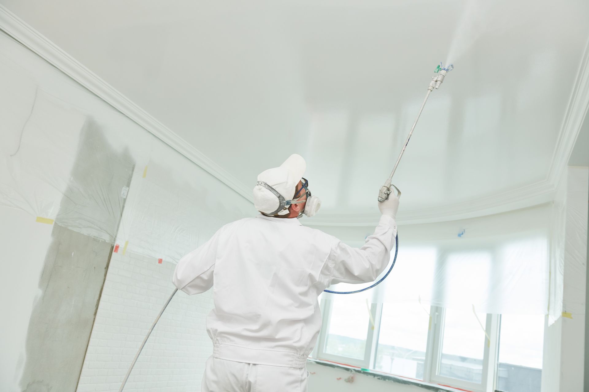 A painter using an airless painting sprayer to cover the ceiling surface with white paint.